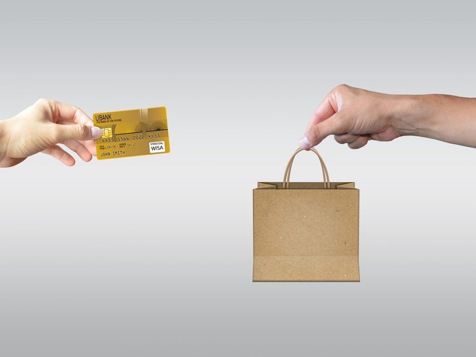 On the left hand holding a credit card reaching out of a computer, on the right hand holding a shopping bag reaching out of another computer. Symbolizing e-commerce.